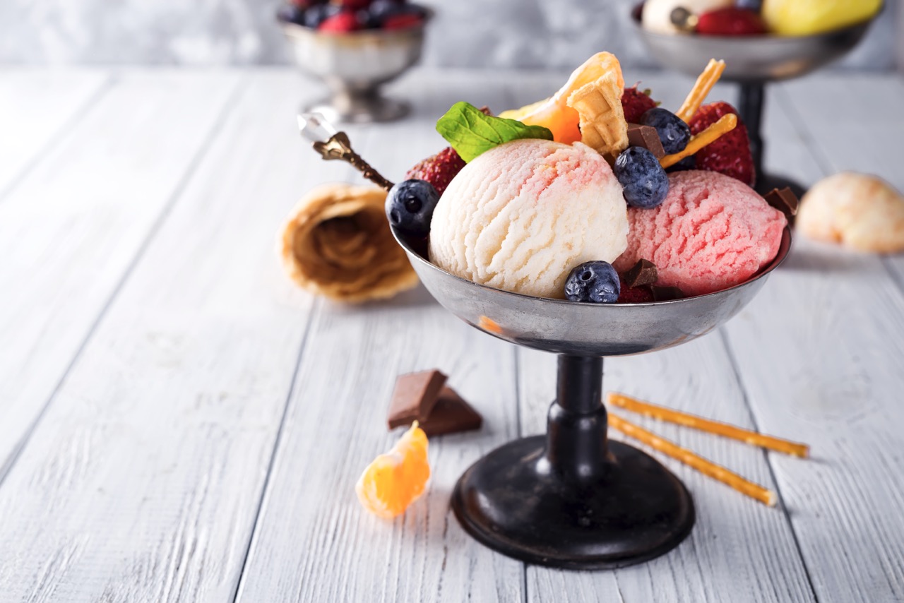 Bowl with ice cream with three different scoops of white, yellow, red colors and waffle cone, chocolate, tangerines and straws