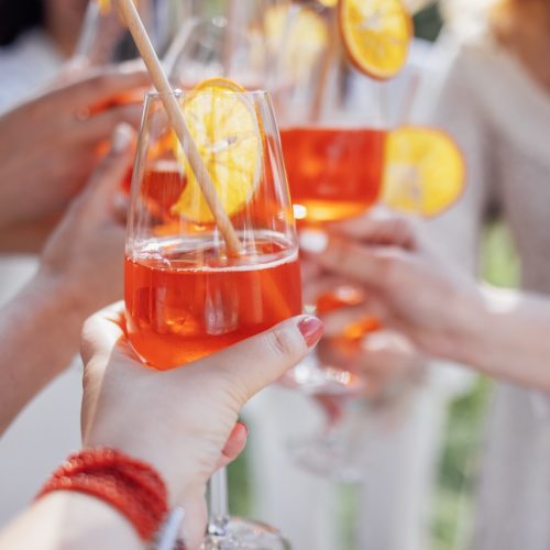 Close-up of a female hands with glasses with a delicious aperol. Young women in white clothes make toast with orange alcoholic drinks. Elegant girls clink cups at a party outdoors.